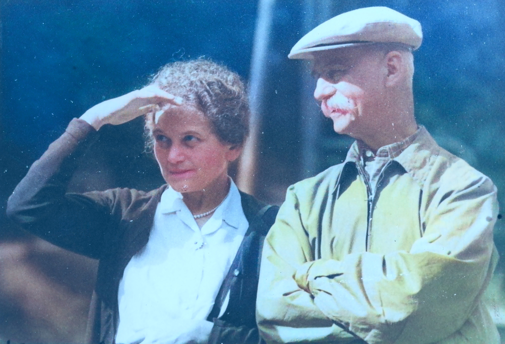 Na zdjęciu widać mężczyznę i kobietę stojących i patrzących przed siebie. Kobieta trzyma prawą dłoń nad czołem i przysłania oczy przed ostrym słońcem, którego blask widoczny jest na zdjęciu.