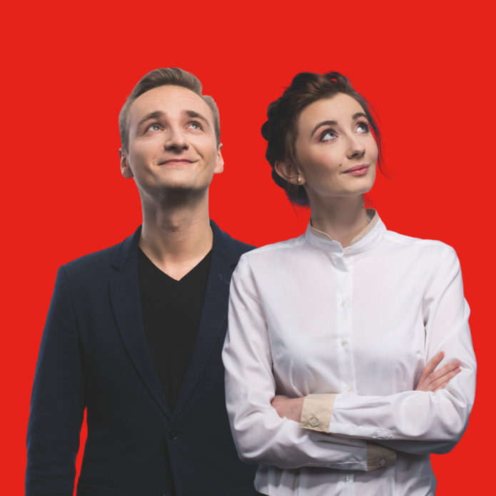 "Młody mężczyzna i kobieta stoją obok siebie na tle intensywnie czerwonego tła, oboje patrzą w górę z lekkimi uśmiechami. Mężczyzna ubrany jest w ciemną marynarkę i czarną koszulkę, a kobieta ma na sobie elegancką, białą koszulę z kołnierzykiem i skrzyżowane ręce."